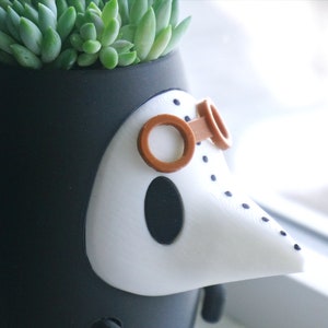 May include: A black and white ceramic planter shaped like a bird with a long beak and brown goggles. The planter has a small succulent plant growing inside.