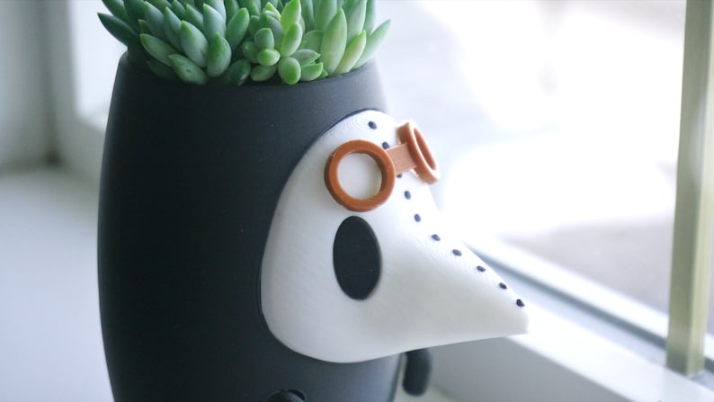 May include: A black and white ceramic planter shaped like a bird with a long beak and brown goggles. The planter has a small succulent plant growing inside.