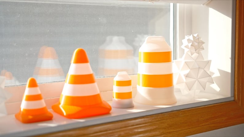 Może przedstawiać: Kolekcja dekoracyjnych przedmiot&oacute;w na parapecie. Przedmioty obejmują pomarańczowo-białe pachołki drogowe o r&oacute;żnych rozmiarach, cylindryczny obiekt z pomarańczowymi paskami i białą geometryczną rzeźbę w kształcie gwiazdy.