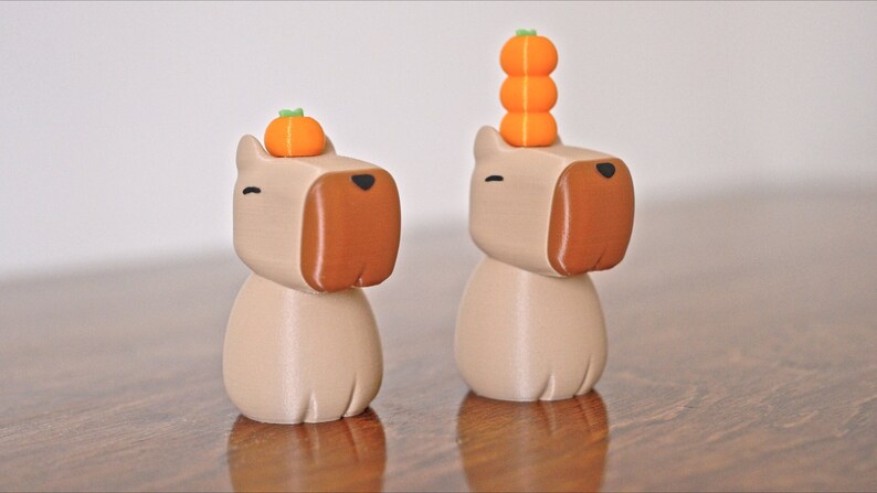 May include: Two beige ceramic dog figurines with brown noses. One dog has a single orange pumpkin on its head, the other has a stack of three orange pumpkins.
