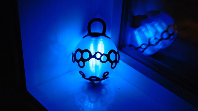 May include: A glowing blue spherical lamp with a black decorative pattern. The lamp has a loop at the top for hanging. The light reflects on the surface below and in the window, creating a bright, ambient glow. The lamp is a decorative lighting fixture.