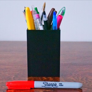 May include: A black pencil holder filled with various pens and pencils, including a yellow highlighter, a blue pen, and a red pen. A red and white Sharpie fine point permanent marker is lying on a wooden surface.