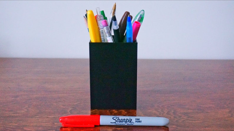 May include: A black pencil holder filled with various pens and pencils, including a yellow highlighter, a blue pen, and a red pen. A red and white Sharpie fine point permanent marker is lying on a wooden surface.