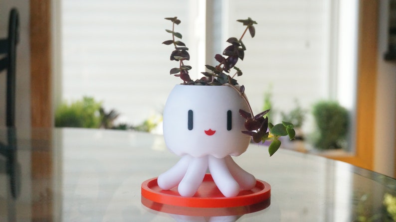 May include: White ceramic planter shaped like an octopus with red lips and black eyes. The planter has five tentacles and is holding a small green plant with red leaves.