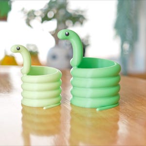 May include: Two green snake-shaped planters sit on a wooden table. The planters are coiled, with a snake head at the top. One is a lighter green than the other. The background is blurred, with a window and plants visible.