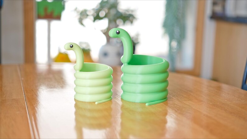 May include: Two green snake-shaped planters sit on a wooden table. The planters are coiled, with a snake head at the top. One is a lighter green than the other. The background is blurred, with a window and plants visible.