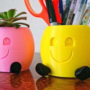 May include: Two 3D printed smiley face planters, one pink with a succulent plant and one yellow with a set of pens inside.