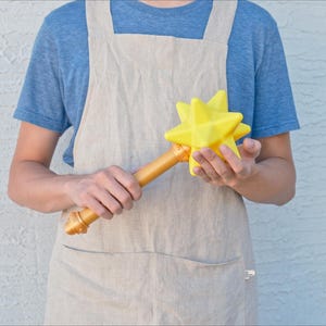 May include: A yellow star-shaped wand with a gold-colored handle. The wand is held by a person wearing a blue shirt and a tan apron. The star has multiple points and a smooth texture.