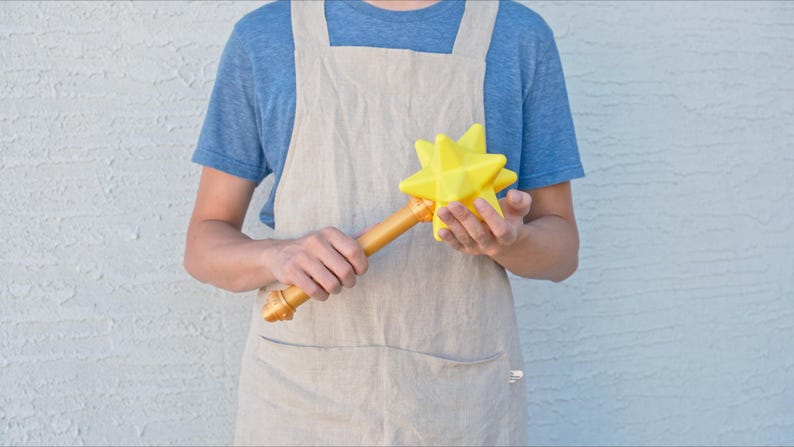 May include: A yellow star-shaped wand with a gold-colored handle. The wand is held by a person wearing a blue shirt and a tan apron. The star has multiple points and a smooth texture.