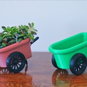 May include: Two miniature wheelbarrows, one red and one green, with black wheels. The red wheelbarrow is filled with a variety of green succulents.