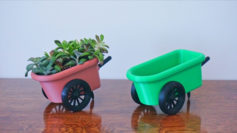 May include: Two miniature wheelbarrows, one red and one green, with black wheels. The red wheelbarrow is filled with a variety of green succulents.