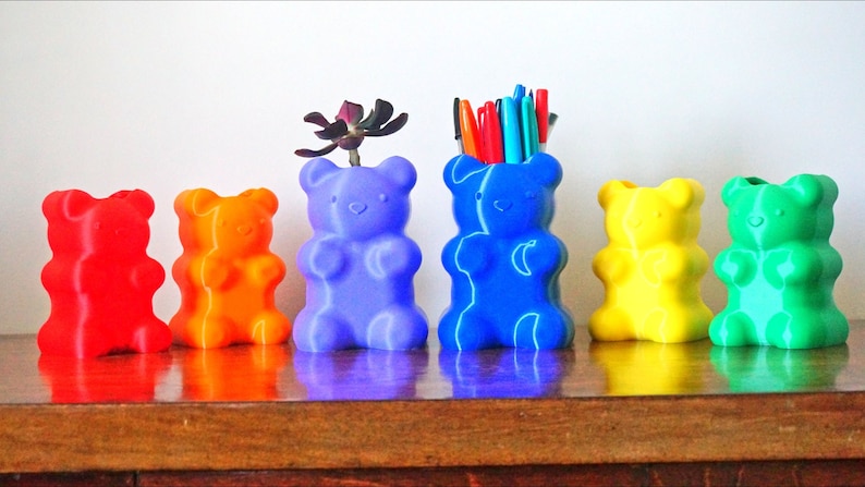 Puede incluir: Seis macetas con forma de osito de goma en un arco&iacute;ris de colores. Las macetas est&aacute;n llenas de plantas y bol&iacute;grafos.