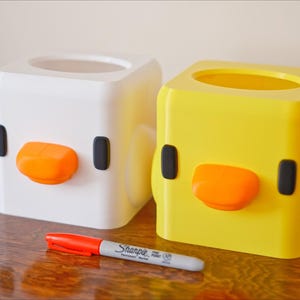 Pu&ograve; includere: Due coperture quadrate per scatola di fazzoletti, una bianca e una gialla, a forma di pulcino dei cartoni animati con occhi neri e becco arancione.