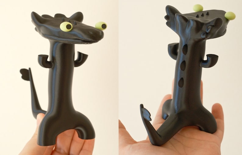 May include: A black, stylized dragon figurine with large, yellow eyes and small green spheres on its head. The dragon has a long neck, a tail, and two small arms. The figurine is held in a hand.