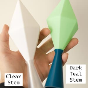May include: Two geometric-shaped objects are held in a hand. One is white with a clear stem, and the other is green with a dark teal stem. The text "Clear Stem" and "Dark Teal Stem" are visible.