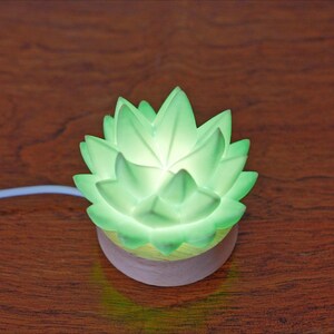 K&ouml;nnte beinhalten: Eine leuchtende gr&uuml;ne Lotusblumen-Nachtlampe. Das Licht hat einen weichen, diffusen Schein und besteht aus einem durchscheinenden Material. Der Sockel ist hellviolett. Ein wei&szlig;es Kabel erstreckt sich von der Basis.