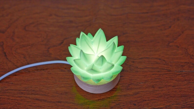 K&ouml;nnte beinhalten: Eine leuchtende gr&uuml;ne Lotusblumen-Nachtlampe. Das Licht hat einen weichen, diffusen Schein und besteht aus einem durchscheinenden Material. Der Sockel ist hellviolett. Ein wei&szlig;es Kabel erstreckt sich von der Basis.
