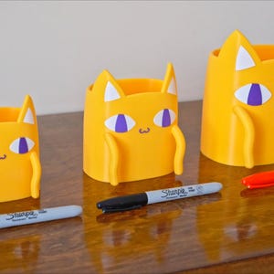 Peut inclure: Trois porte-crayons jaunes en forme de chat de diff&eacute;rentes tailles. Les porte-crayons ont des yeux violets et des oreilles blanches. Le plus grand porte-crayon est &agrave; gauche, le plus petit &agrave; droite. Les porte-crayons sont sur une surface en bois avec trois marqueurs.
