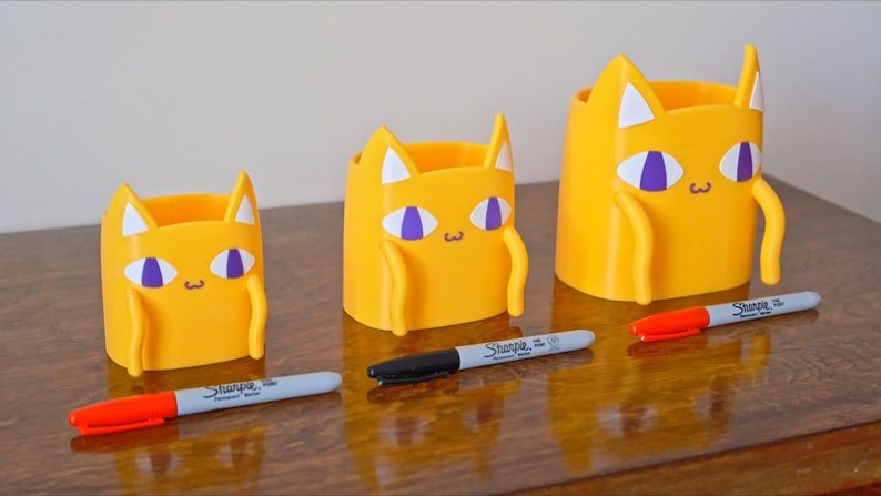 Peut inclure: Trois porte-crayons jaunes en forme de chat de diff&eacute;rentes tailles. Les porte-crayons ont des yeux violets et des oreilles blanches. Le plus grand porte-crayon est &agrave; gauche, le plus petit &agrave; droite. Les porte-crayons sont sur une surface en bois avec trois marqueurs.
