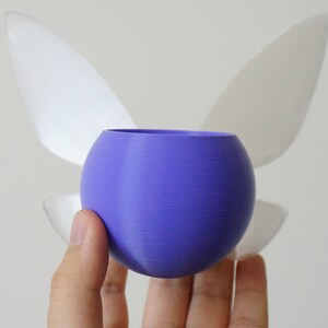 Puede incluir: Una esfera p&uacute;rpura con dos alas blancas unidas a la parte posterior. La esfera est&aacute; siendo sostenida en una mano.