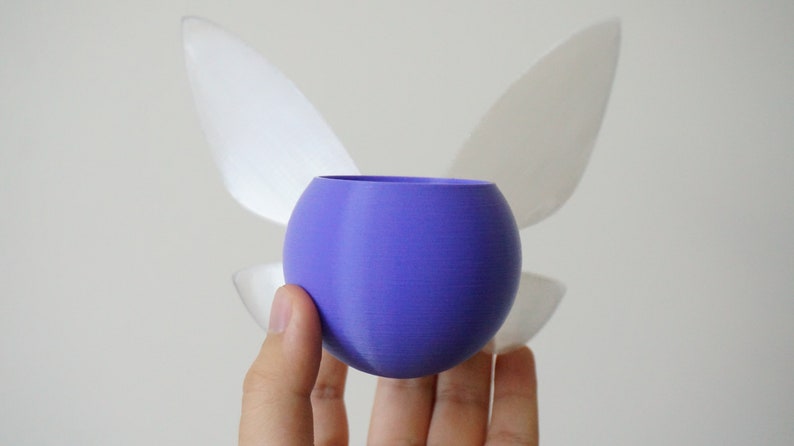 Puede incluir: Una esfera p&uacute;rpura con dos alas blancas unidas a la parte posterior. La esfera est&aacute; siendo sostenida en una mano.