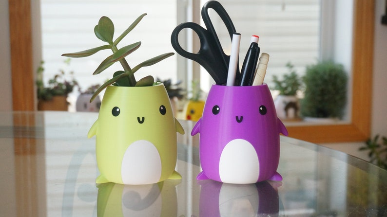 May include: Two cute penguin-shaped planters, one yellow with a green succulent plant and the other purple with a white and black pen holder.
