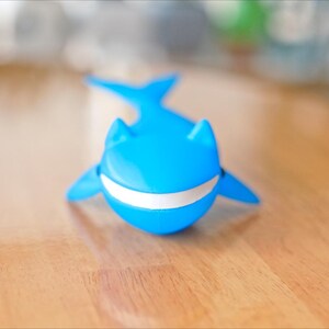 May include: A bright blue whale-shaped toy with a white stripe across its mouth. The toy has fins and a tail, and is sitting on a wooden surface. The toy is a fun and playful item.