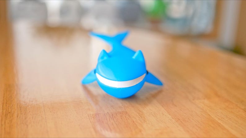 May include: A bright blue whale-shaped toy with a white stripe across its mouth. The toy has fins and a tail, and is sitting on a wooden surface. The toy is a fun and playful item.