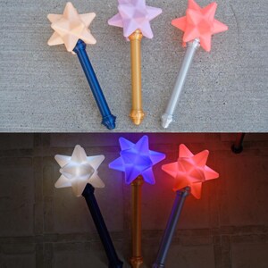 May include: A collection of star-shaped wands in various colors, including peach, lavender, and coral, with gold, silver, and blue handles. The wands have a light-up feature, with the stars glowing in white, blue, and red.