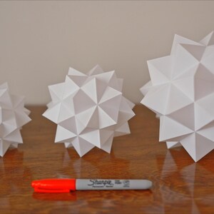 Pode incluir: Tr&ecirc;s formas geom&eacute;tricas de papel branco de tamanhos diferentes. As formas s&atilde;o todas feitas de papel dobrado e t&ecirc;m um design de estrela. As formas est&atilde;o dispostas em uma fila da menor para a maior.