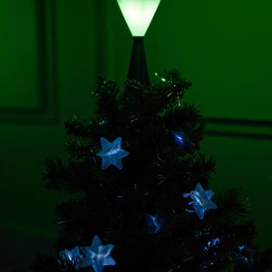 May include: A small, dark green Christmas tree with blue star-shaped lights and a bright green diamond-shaped tree topper. The tree is set against a dark green background, creating a festive holiday scene.