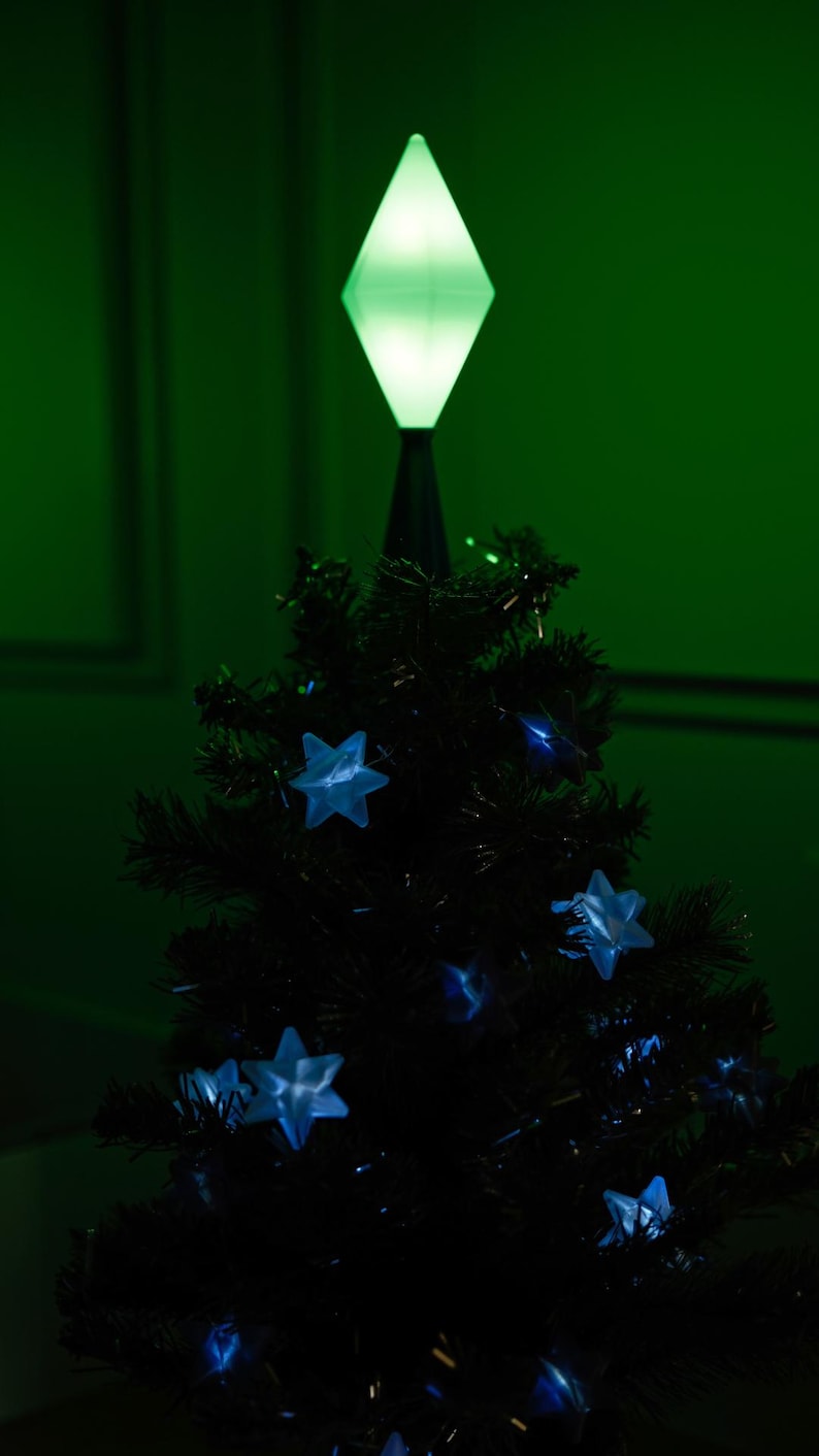 May include: A small, dark green Christmas tree with blue star-shaped lights and a bright green diamond-shaped tree topper. The tree is set against a dark green background, creating a festive holiday scene.