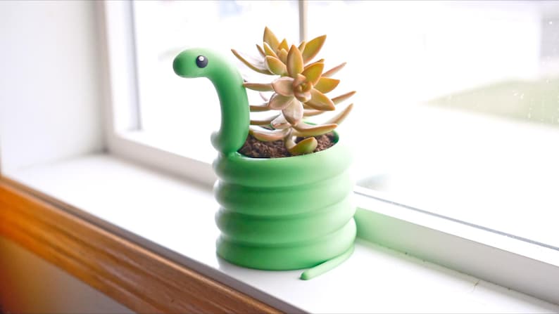 May include: A green snake-shaped planter with a succulent plant. The planter has a coiled body and a snake head that curves upwards. The succulent has green and yellow-orange leaves. The planter is on a windowsill.