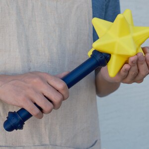 May include: A yellow star-shaped wand with a dark blue handle. The star has multiple points and is held in the hands. The handle has decorative details. The wand is a toy or prop.