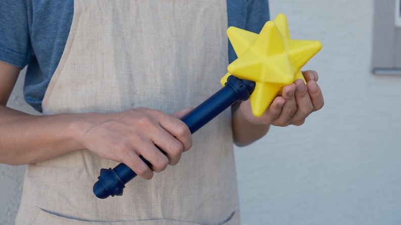 May include: A yellow star-shaped wand with a dark blue handle. The star has multiple points and is held in the hands. The handle has decorative details. The wand is a toy or prop.