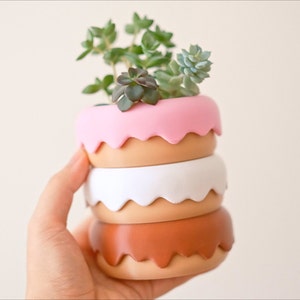 Puede incluir: Un conjunto de cuatro macetas apiladas en forma de donut en rosa, marr&oacute;n, blanco y marr&oacute;n. La maceta superior tiene una planta suculenta creciendo en ella.