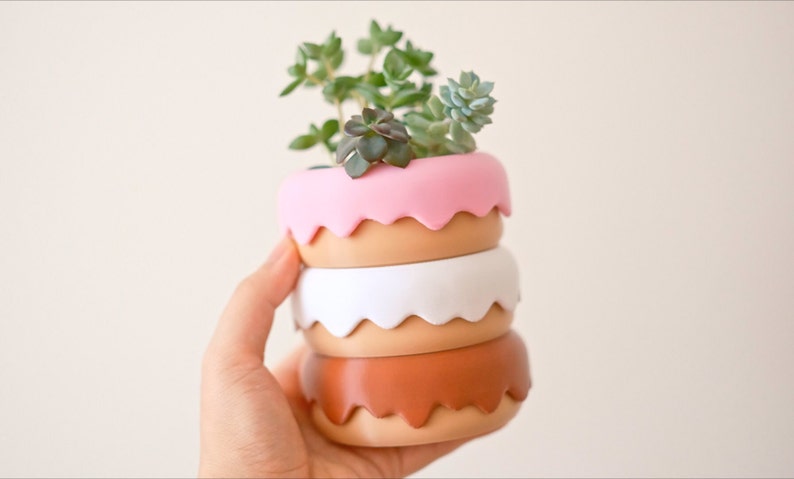 Puede incluir: Un conjunto de cuatro macetas apiladas en forma de donut en rosa, marr&oacute;n, blanco y marr&oacute;n. La maceta superior tiene una planta suculenta creciendo en ella.