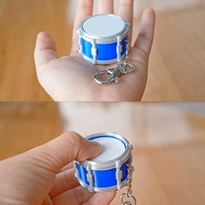 May include: A miniature drum keychain with a blue body, silver accents, and a white drumhead. The keychain includes a silver chain and clasp. The drum is held in a hand, showcasing its small size and detailed design.