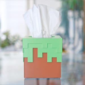 A cube tissue box cover inspired by the dirt grass block from the popular video game, Minecraft, is on a kitchen table with plants in the background. It has bright green pixelated grass at the top half and the bottom half is brown.