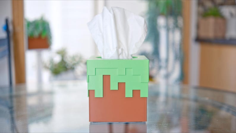 A cube tissue box cover inspired by the dirt grass block from the popular video game, Minecraft, is on a kitchen table with plants in the background. It has bright green pixelated grass at the top half and the bottom half is brown.