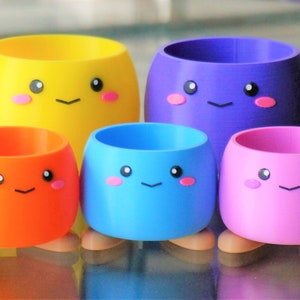 May include: Set of four colorful, small, round planters with cute faces. The planters are yellow, orange, blue, and pink. Each planter has two small feet.