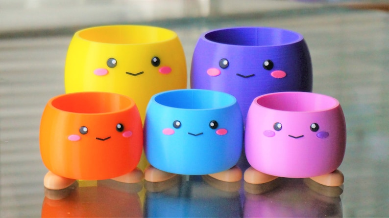 May include: Set of four colorful, small, round planters with cute faces. The planters are yellow, orange, blue, and pink. Each planter has two small feet.