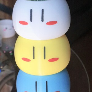 May include: A stack of four colorful ceramic planters shaped like Japanese dango, a sweet dumpling. The planters are white, yellow, blue, and pink. Each planter has a simple black face with two eyes and a mouth. The top planter has a small succulent plant growing in it.