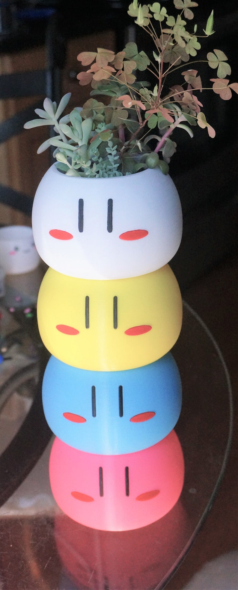 May include: A stack of four colorful ceramic planters shaped like Japanese dango, a sweet dumpling. The planters are white, yellow, blue, and pink. Each planter has a simple black face with two eyes and a mouth. The top planter has a small succulent plant growing in it.