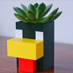 May include: A black, white, yellow, and red planter shaped like a pixelated face with a green succulent plant growing inside.