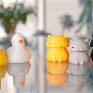 Peut inclure: Cinq petites figurines d'animaux imprim&eacute;es en 3D. Un canard jaune, un oiseau blanc, un chat orange, un chat gris et un chat noir. Les figurines sont toutes assises sur une surface r&eacute;fl&eacute;chissante.