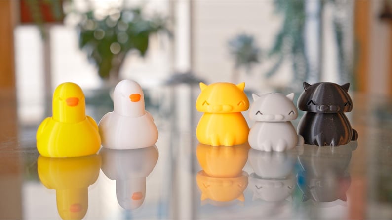 Peut inclure: Cinq petites figurines d'animaux imprim&eacute;es en 3D. Un canard jaune, un oiseau blanc, un chat orange, un chat gris et un chat noir. Les figurines sont toutes assises sur une surface r&eacute;fl&eacute;chissante.