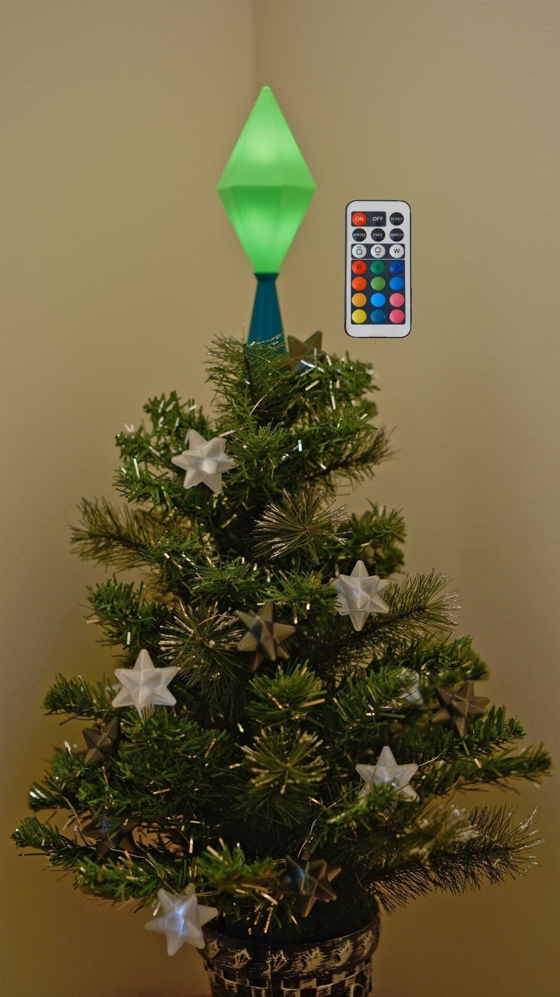 May include: A small artificial Christmas tree with star ornaments and a green, diamond-shaped tree topper. A remote control with color options is included. The tree is decorated with silver tinsel and star-shaped ornaments.