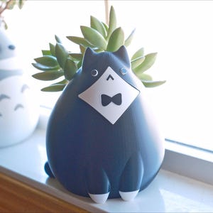 May include: A navy blue cat-shaped planter with a white diamond-shaped bib and bow tie. The planter holds a succulent plant. Another gray planter in the shape of a cartoon character is visible in the background. The planters are on a windowsill.