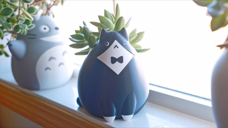May include: A navy blue cat-shaped planter with a white diamond-shaped bib and bow tie. The planter holds a succulent plant. Another gray planter in the shape of a cartoon character is visible in the background. The planters are on a windowsill.